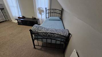 Une petite chambre à coucher, bien rangée, avec un lit simple, une table de chevet, un ventilateur sur pied et une fenêtre avec des rideaux.