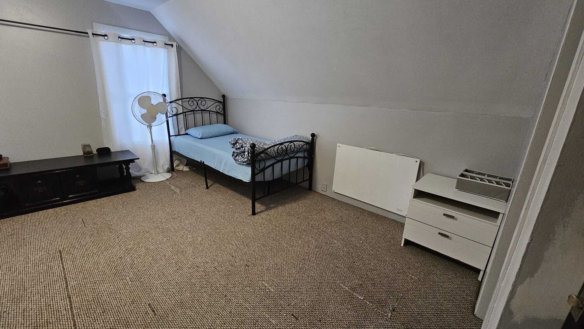 Quarto pequeno com cama de solteiro, teto inclinado, mesa de cabeceira, ventoinha, cômoda e radiador.