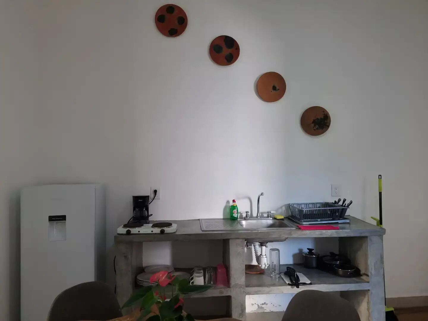 A compact kitchen with a sink, a coffee maker, and a small refrigerator. There are decorative plates on the wall and a plant on the counter.