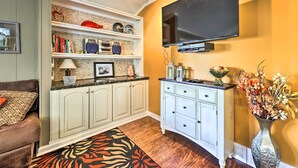 Un salon avec un téléviseur fixé au mur, un meuble blanc avec des tiroirs, un canapé agrémenté de coussins à motifs et un vase décoratif avec des fleurs.