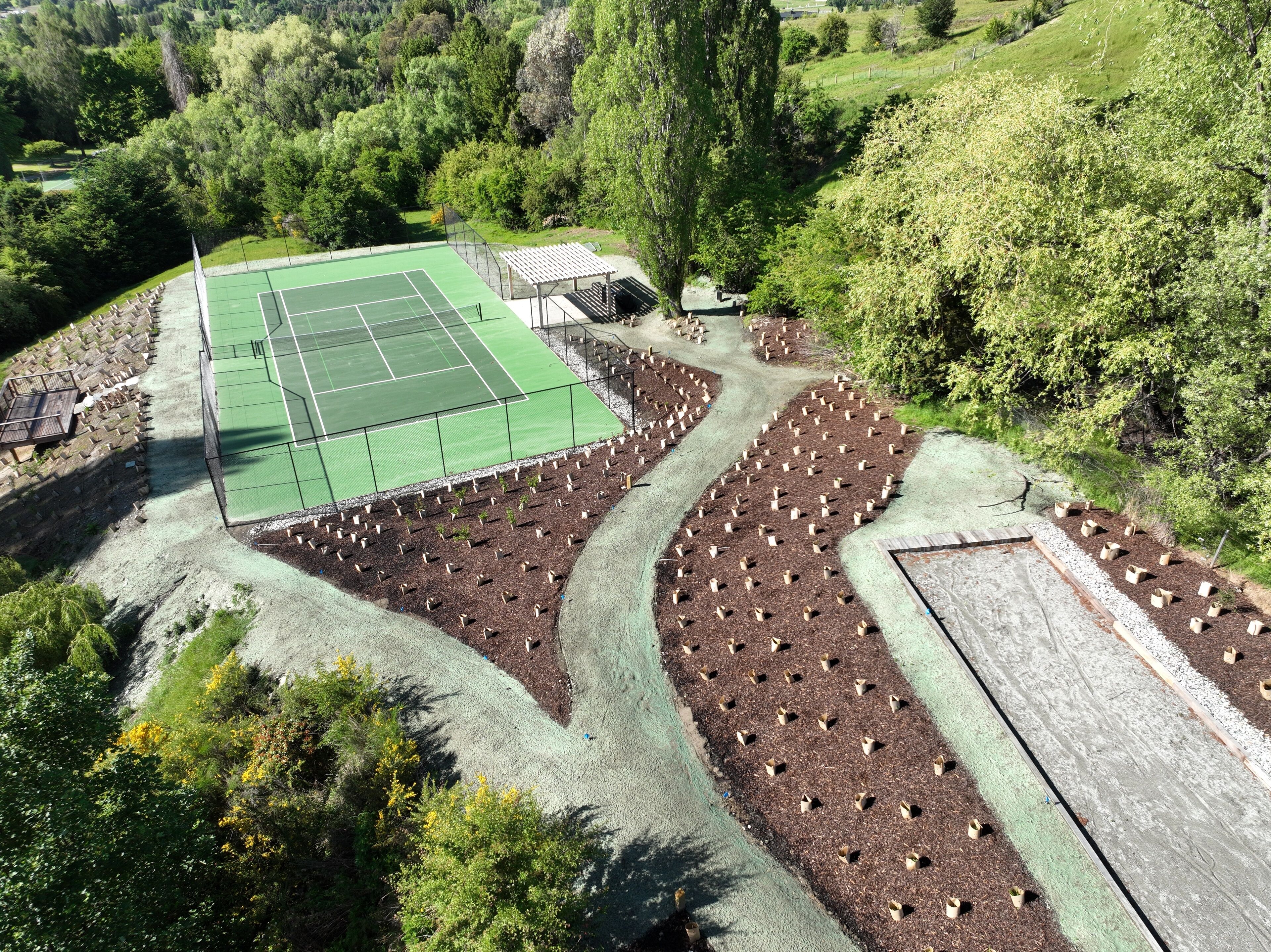 An aerial view of a tennis court surrounded by landscaped areas with young trees and shrubs.