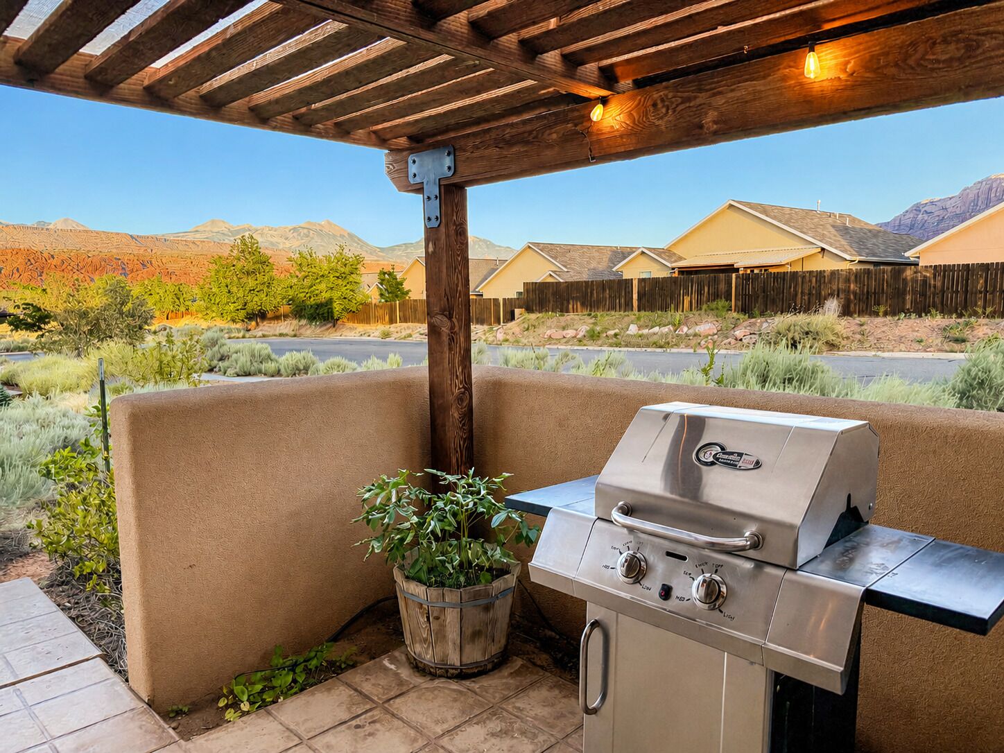 Une terrasse extérieure dotée d’un barbecue intégré, d’une pergola en bois au-dessus et offrant une vue sur un quartier résidentiel, avec des maisons et des montagnes en arrière-plan.