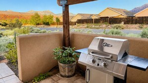 An outdoor patio with a built-in grill, a wooden pergola overhead, and a view of a residential area with houses and mountains in the background.