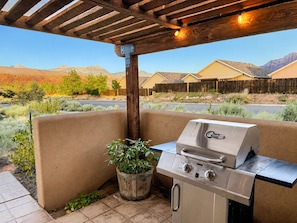 An outdoor patio with a built-in grill, a wooden pergola overhead, and a view of a residential area with houses and mountains in the background.