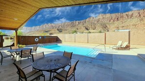 An outdoor pool area with a wooden pergola, a round table with chairs, and a view of a rocky hillside.