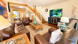A spacious living room with a wooden staircase, leather sofas, a coffee table, and a flat-screen TV.