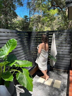 A person in a towel at a shower area with a wooden fence and large green plants. - Custom Designed Tiny Home (Ulladulla)