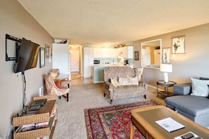 Un salon avec un canapé, un fauteuil et une table basse, une cuisine à l’arrière-plan et un téléviseur fixé au mur.