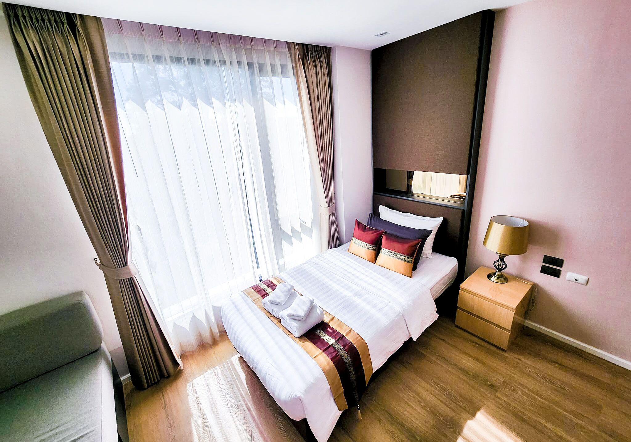 A hotel room with a large bed, a bedside table with a lamp, and a view of the outdoors through sheer curtains.
