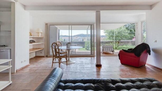 Ein geräumiger Wohnbereich mit einer Couch, einem Esstisch mit Stühlen, einem roten Sessel und Blick durch eine Glastür.