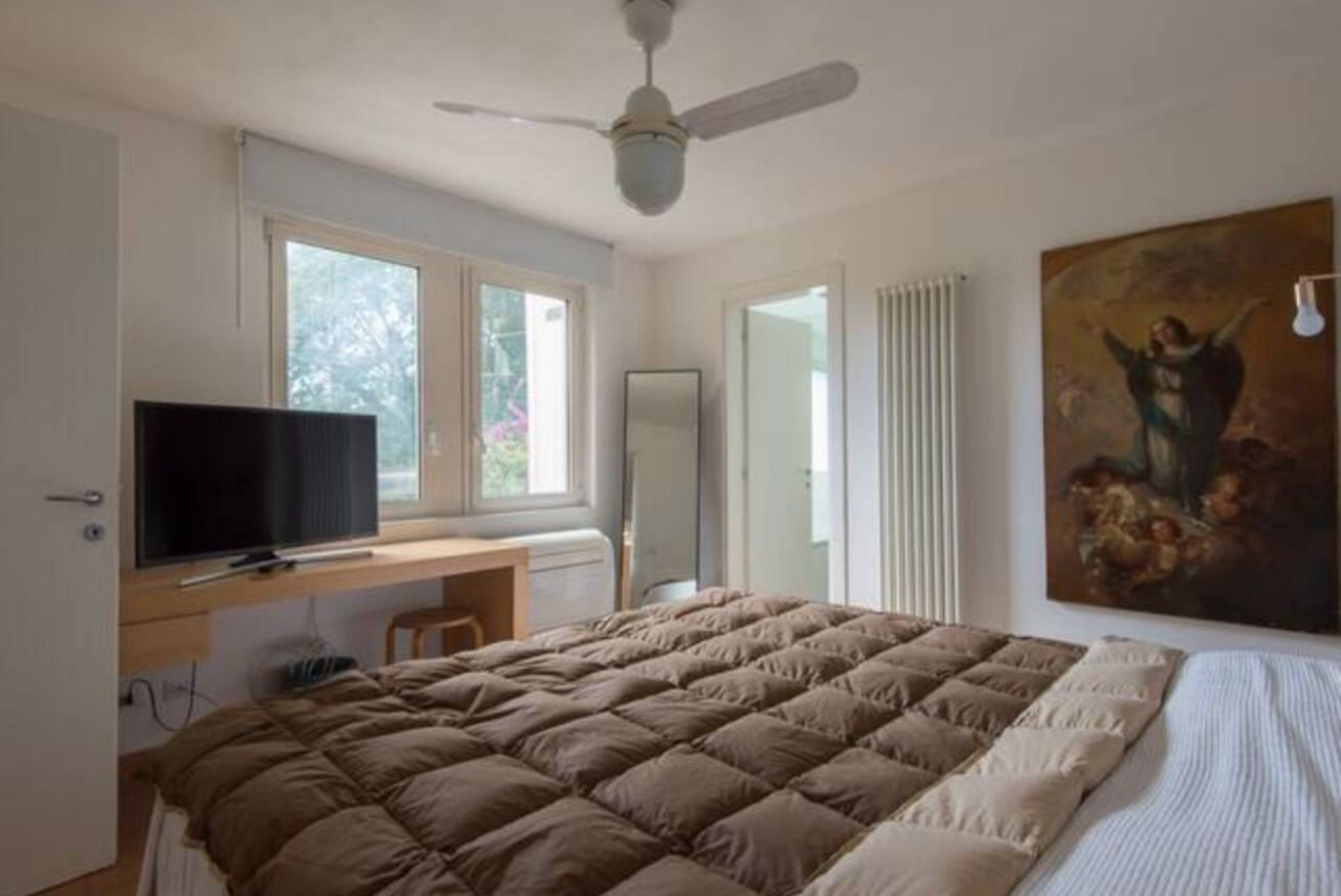 A bedroom with a bed, a desk, a television, a ceiling fan, and a painting.