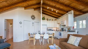 Un salón espacioso con techo de madera, una mesa de comedor con sillas, un sofá y una zona de cocina con armarios y electrodomésticos blancos.