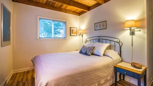 Un dormitorio con cama, vigas de madera en el techo, una ventana con persianas, dos lámparas de pared y una mesita de noche con lámpara.