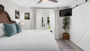 Un dormitorio con una cama, ventilador de techo, televisor, planta y puerta corrediza a un balcón.