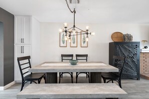 A modern dining area with a large rectangular table, two chairs, a bowl on the table, a chandelier, and a decorative wall panel.