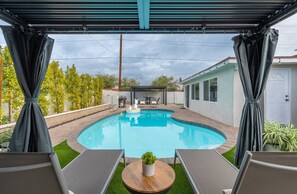 A backyard with a swimming pool, a covered patio area, and a small table with a plant.