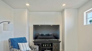 A modern living room with a flat-screen TV mounted on a black stand, a patterned armchair with a blue throw, and a floor lamp. - Ideal Family Getaway- Pickleball, Pool & Near Zion (Washington)