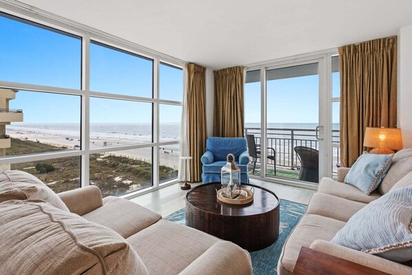 A spacious living room with large windows, a blue armchair, a beige sofa, and a round coffee table.