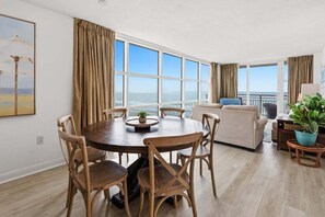 A spacious living area with a round wooden table, beige sofa, and large windows offering ocean views.