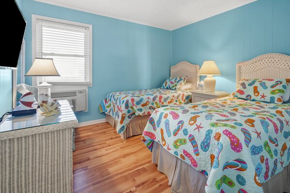 A bedroom with two beds, a wicker chair, a table with a lamp, and a window with blinds.