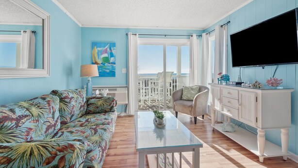 A room with a blue wall, a white sofa, a glass coffee table, a white cabinet, and a TV. - Stunning views, stylish oceanfront condo close to nightlife, shopping, dining and more! (North Myrtle Beach)