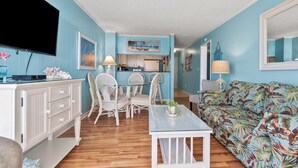 A living room with a television, a dining table with chairs, a sofa, and a kitchen area in the background. - Stunning views, stylish oceanfront condo close to nightlife, shopping, dining and more! (North Myrtle Beach)
