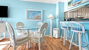A room with a round glass table surrounded by wicker chairs, a white floor lamp, and a kitchen area with a blue wall and a painting above the counter. - Stunning views, stylish oceanfront condo close to nightlife, shopping, dining and more! (North Myrtle Beach)
