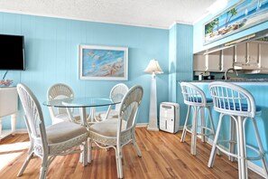 A room with a round glass table surrounded by wicker chairs, a white floor lamp, and a kitchen area with a blue wall and a painting above the counter.