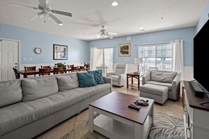 A spacious living room with a grey sofa, a coffee table, and a TV stand. There are two ceiling fans, a dining area with wooden chairs, and a large window with curtains. - Spacious 5 bedroom oceanview condo - Cherry Grove Beach (North Myrtle Beach)