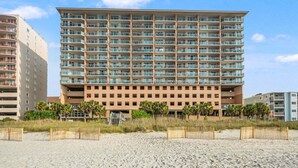 Un hôtel à plusieurs étages doté de balcons, situé en bord de plage, avec une plage de sable devant et un ciel dégagé.