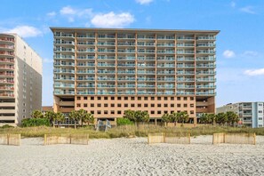 A multi-story hotel with balconies, a sandy beach in front, and a clear sky.