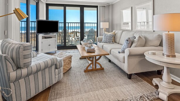Uma sala de estar moderna com uma TV grande, um sofá, uma mesa de centro e uma varanda com vista para a praia.