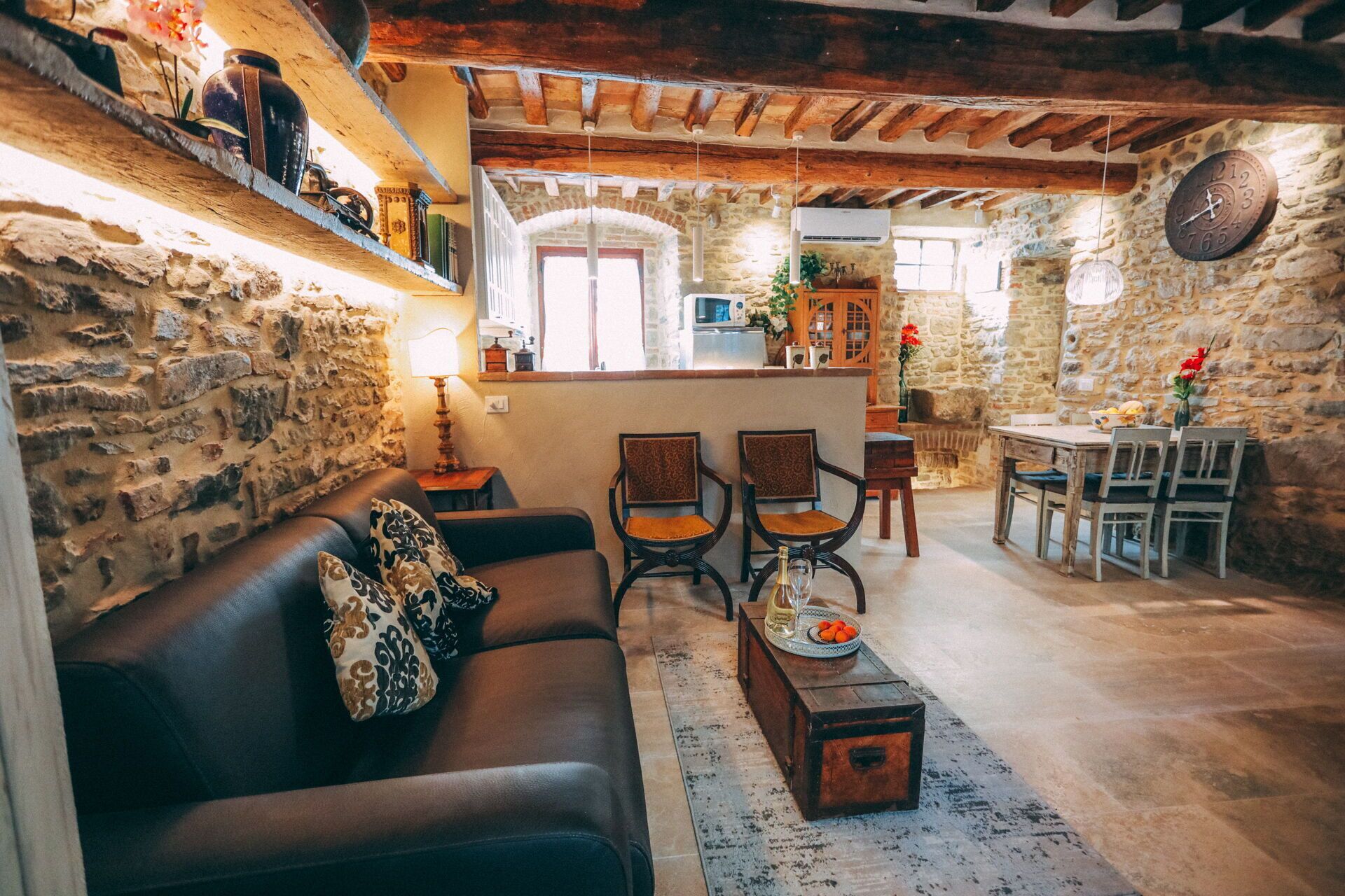 Un interno rustico con parete in pietra, travi in legno a soffitto, un divano in pelle, due poltrone, un tavolino in legno e un'area pranzo con tavolo e sedie.