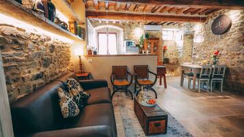 Un interno rustico con parete in pietra, travi in legno a soffitto, un divano in pelle, due poltrone, un tavolino in legno e un'area pranzo con tavolo e sedie.