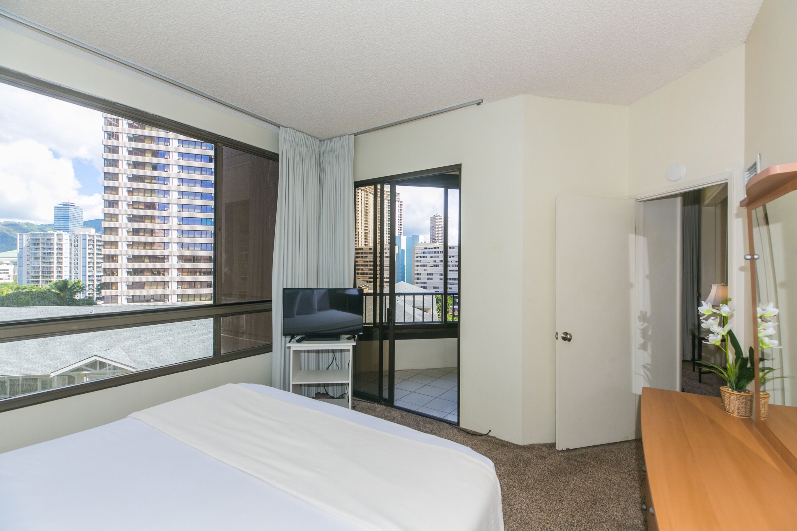 Une chambre d’hôtel avec un lit, une télévision, un balcon donnant sur la ville et une commode en bois.