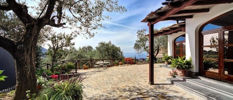 A stone-paved outdoor area with a pergola, potted plants, and a view of the sea.