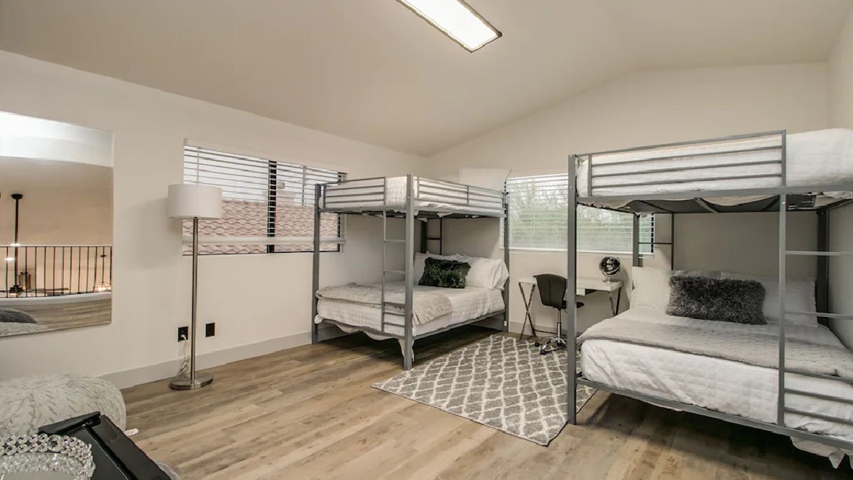 A dormitory room with bunk beds, a desk, a chair, a lamp, and a patterned rug.