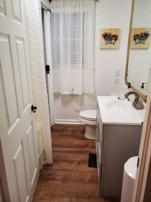 Une salle de bain avec des toilettes blanches, un lavabo doté d’un robinet doré et un rideau de douche. Un tableau est accroché au mur et un miroir se trouve au-dessus du lavabo.