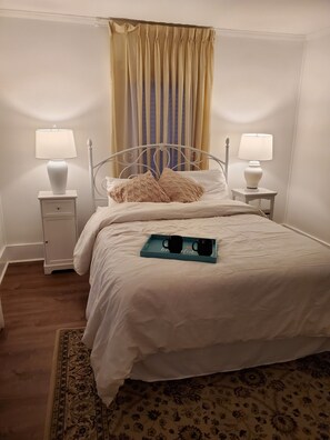 Un lit bien fait, avec du linge de lit blanc, deux tables de chevet avec des lampes, un plateau bleu avec deux tasses noires et un tapis à motifs sur un plancher en bois.
