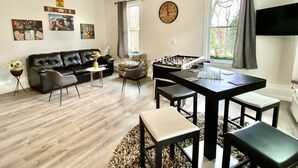 A modern living room with a black leather sofa, a small round table, a foosball table, and a flat-screen TV mounted on the wall. - Renovated Ranch Home. Pet Friendly, Game Room, Fire Pit, Grill, Covered Porch (Woodstock)