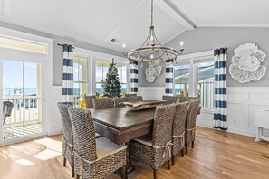 A dining area with a large wooden table, wicker chairs, a chandelier, and a view of the beach through large windows.