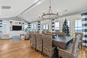 Un vaste espace dédié à la salle à manger, avec une grande table en bois et des chaises en osier, un lustre, et une vue sur l’océan grâce à de grandes fenêtres.