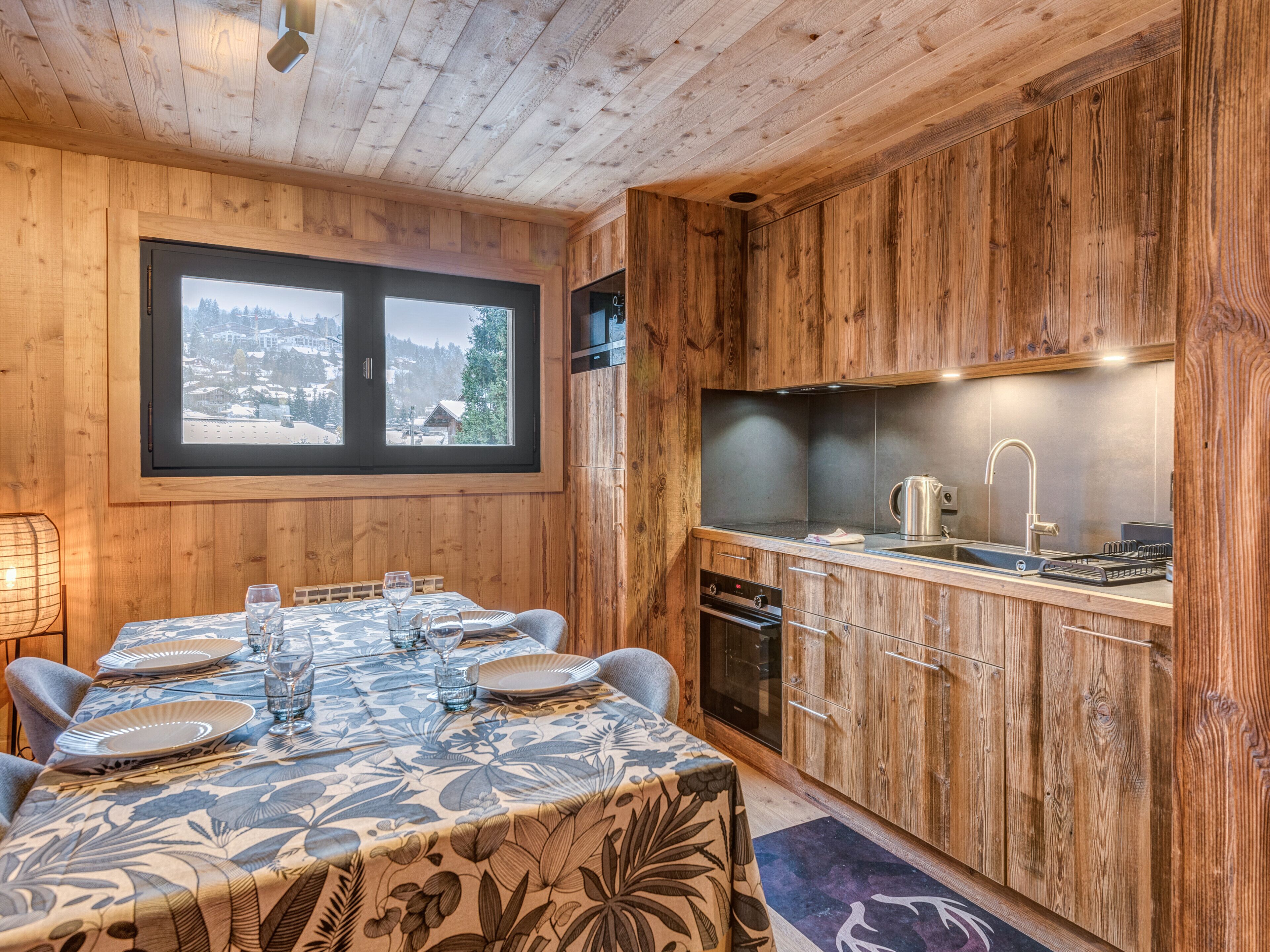 A wooden interior with a dining table set for six, a kitchen with wooden cabinets, and a window overlooking a snowy landscape.
