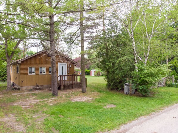 Une cabane en bois avec un porche, entourée d’arbres et d’un petit abri de jardin.