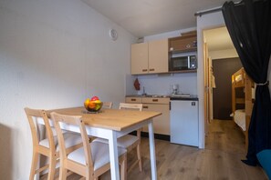 A compact kitchen with wooden cabinets, a microwave, and a small refrigerator. There is a dining table with chairs and a fruit bowl on it. - Studio Belledonne - Welkeys (Huez)