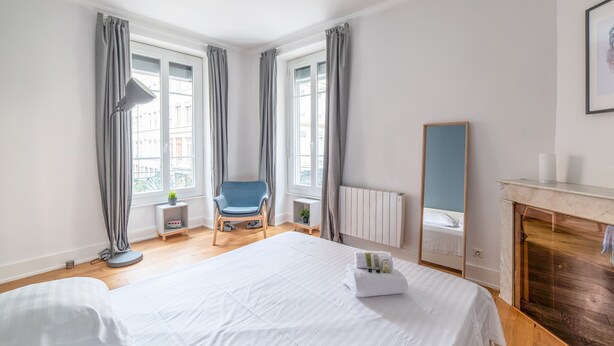A bedroom with a large bed, a chair, a radiator, a fireplace, and a window with curtains. - Appartement Anatha - Welkeys (Lyon)