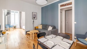 A modern living room with a grey sofa, a dining table set for four, and a large window with curtains. - Appartement Anatha - Welkeys (Lyon)