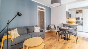 A modern living room with a grey sofa, a small wooden table, and a dining area with chairs. - Appartement Anatha - Welkeys (Lyon)