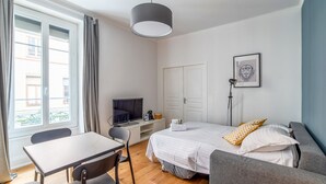 A modern bedroom with a bed, a desk, a chair, a sofa, and a window with curtains. - Appartement Anatha - Welkeys (Lyon)