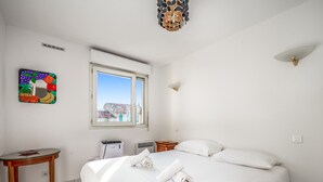 A hotel room with a large bed, a small table, a heater, and a window with a view of buildings. - Appartement California - Welkeys (Nice)